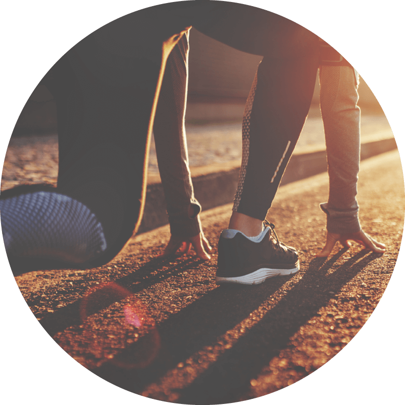 Person in Startposition hockend, mit Sportschuhen, auf einer sonnenbeschienenen Straße, die sich auf das Laufen vorbereitet.
