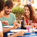 Drei Personen lächeln und essen gemeinsam an einem Tisch im Freien, umgeben von Getränken, Flaschen und verschwommenem Grün.