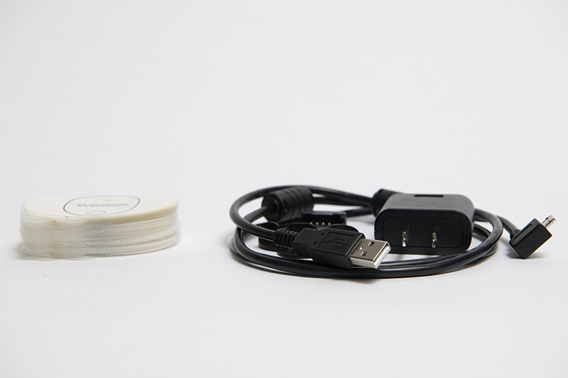 Ein USB-Ladekabel mit Adapter liegt aufgerollt auf einer ebenen Fläche neben den Transmitterpflastern.
